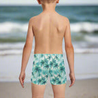 Child wearing tropical palm tree patterned swim shorts standing on a beach and looking out over the ocean
