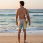 Guy wearing Pastel striped shorts on the beach is looking out at the ocean holding a surfboard 