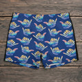 Blue swim shorts with colorful hang loose shaka signs pattern on a beach.