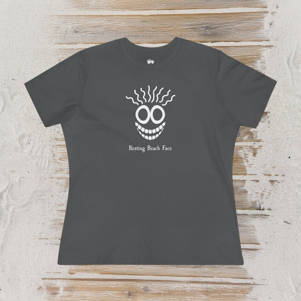 A dark gray short-sleeve t-shirt with a white graphic of a skull-like face with wavy hair and the text 'Resting Beach Face' printed on the chest, displayed flat on a light wood plank background.