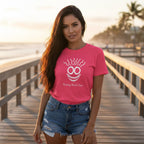 A bright pink short-sleeve t-shirt with a white graphic of a smiling face with wavy hair and the text 'Renting Beach Face' printed on the chest, displayed flat on a light wood plank background.