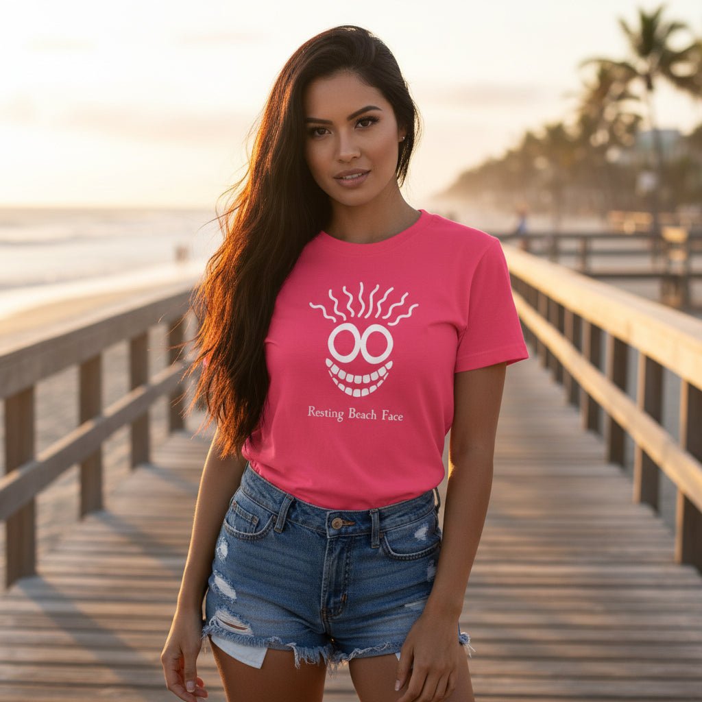 A bright pink short-sleeve t-shirt with a white graphic of a smiling face with wavy hair and the text 'Renting Beach Face' printed on the chest, displayed flat on a light wood plank background.