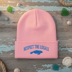 A light pink ribbed knit beanie hat with a folded cuff, featuring blue embroidered text reading 'RESPECT THE LOCALS' and a blue manatee graphic on the front, set against a plain white background.