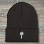 A dark charcoal gray knit beanie with a folded cuff, featuring a small white embroidered palm tree on the front, displayed flat on a rustic, weathered wood background.