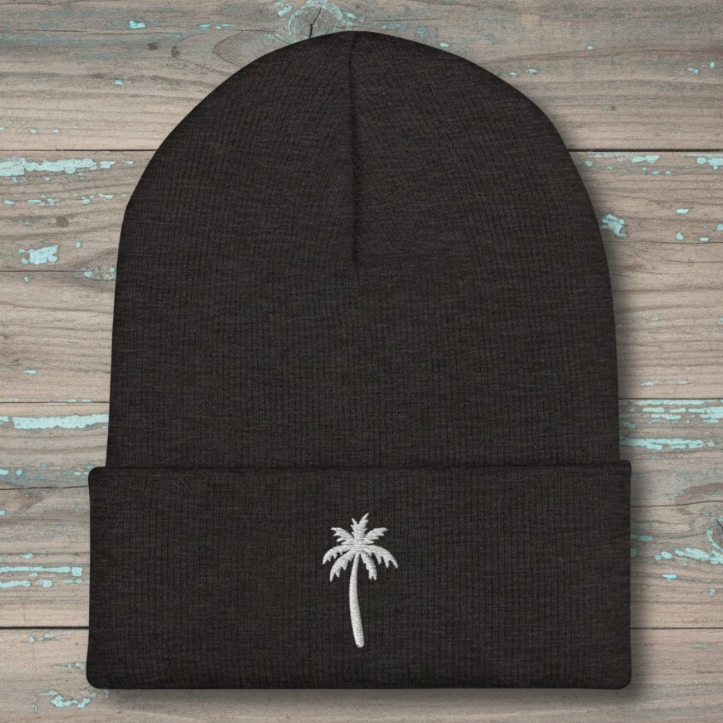 A dark charcoal gray knit beanie with a folded cuff, featuring a small white embroidered palm tree on the front, displayed flat on a rustic, weathered wood background.
