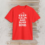 A bright red short-sleeve t-shirt with a white graphic print reading 'KEEP CALM AND SURF BOMBS' and a wave icon, displayed flat on a light wood plank background.