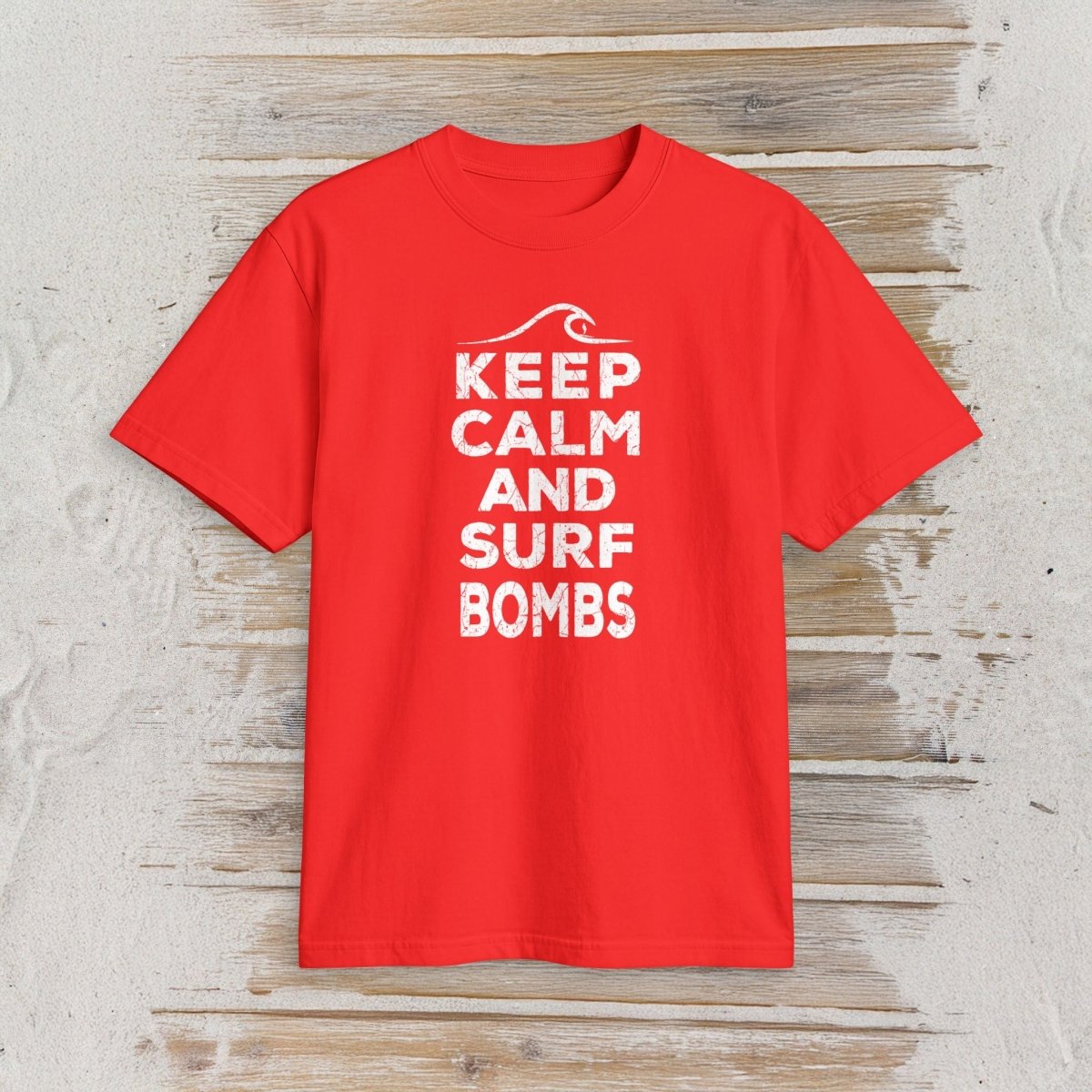 A bright red short-sleeve t-shirt with a white graphic print reading 'KEEP CALM AND SURF BOMBS' and a wave icon, displayed flat on a light wood plank background.