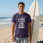 A man standing on a sandy beach holding a white surfboard, wearing a dark purple t-shirt with the white text 'KEEP CALM AND SURF BOMBS' and light blue jeans.