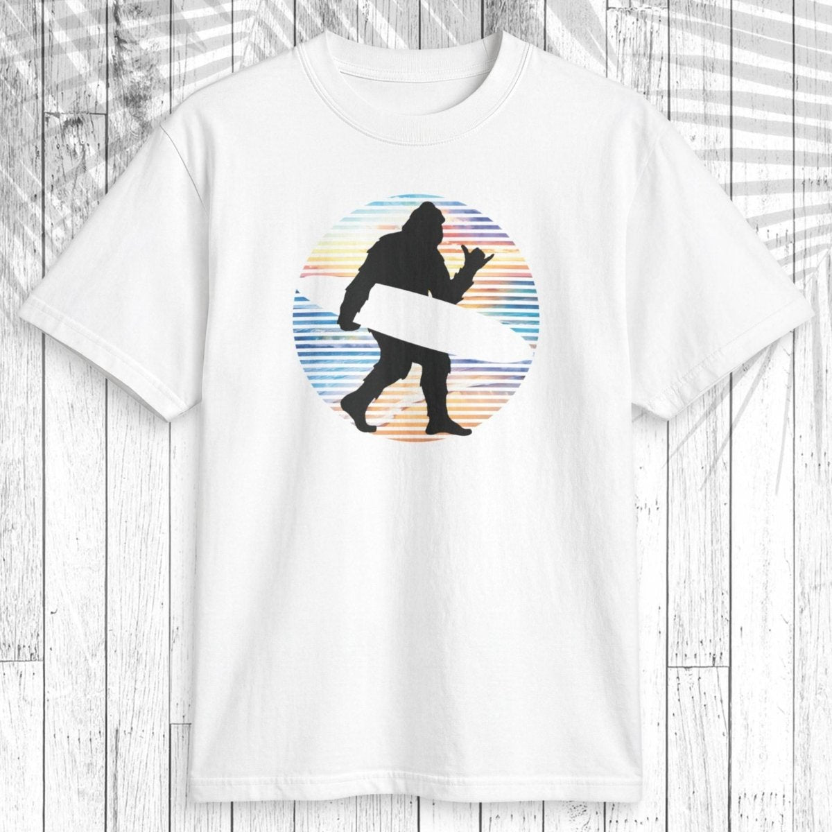 A white short-sleeve crewneck t-shirt featuring a circular graphic of a black silhouette of Bigfoot carrying a white surfboard, set against a background of horizontal pastel blue, orange, and yellow stripes. The shirt is displayed flat on a white wood plank background.