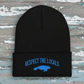 A black ribbed knit beanie with a folded cuff featuring blue embroidered text reading 'RESPECT THE LOCALS' above a blue manatee graphic, shown on a rustic wood