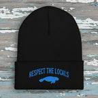 A black ribbed knit beanie with a folded cuff featuring blue embroidered text reading 'RESPECT THE LOCALS' above a blue manatee graphic, shown on a rustic wood