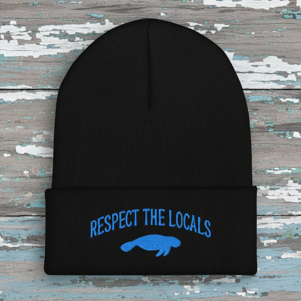 A black ribbed knit beanie with a folded cuff featuring blue embroidered text reading 'RESPECT THE LOCALS' above a blue manatee graphic, shown on a rustic wood