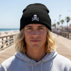 A black knit cuffed beanie hat featuring an embroidered white skull and crossbones design on the front cuff, shown against a plain white background.
