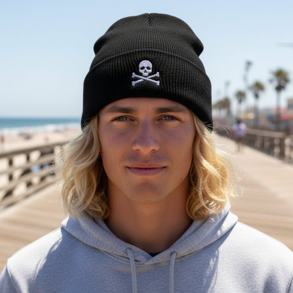 A black knit cuffed beanie hat featuring an embroidered white skull and crossbones design on the front cuff, shown against a plain white background.