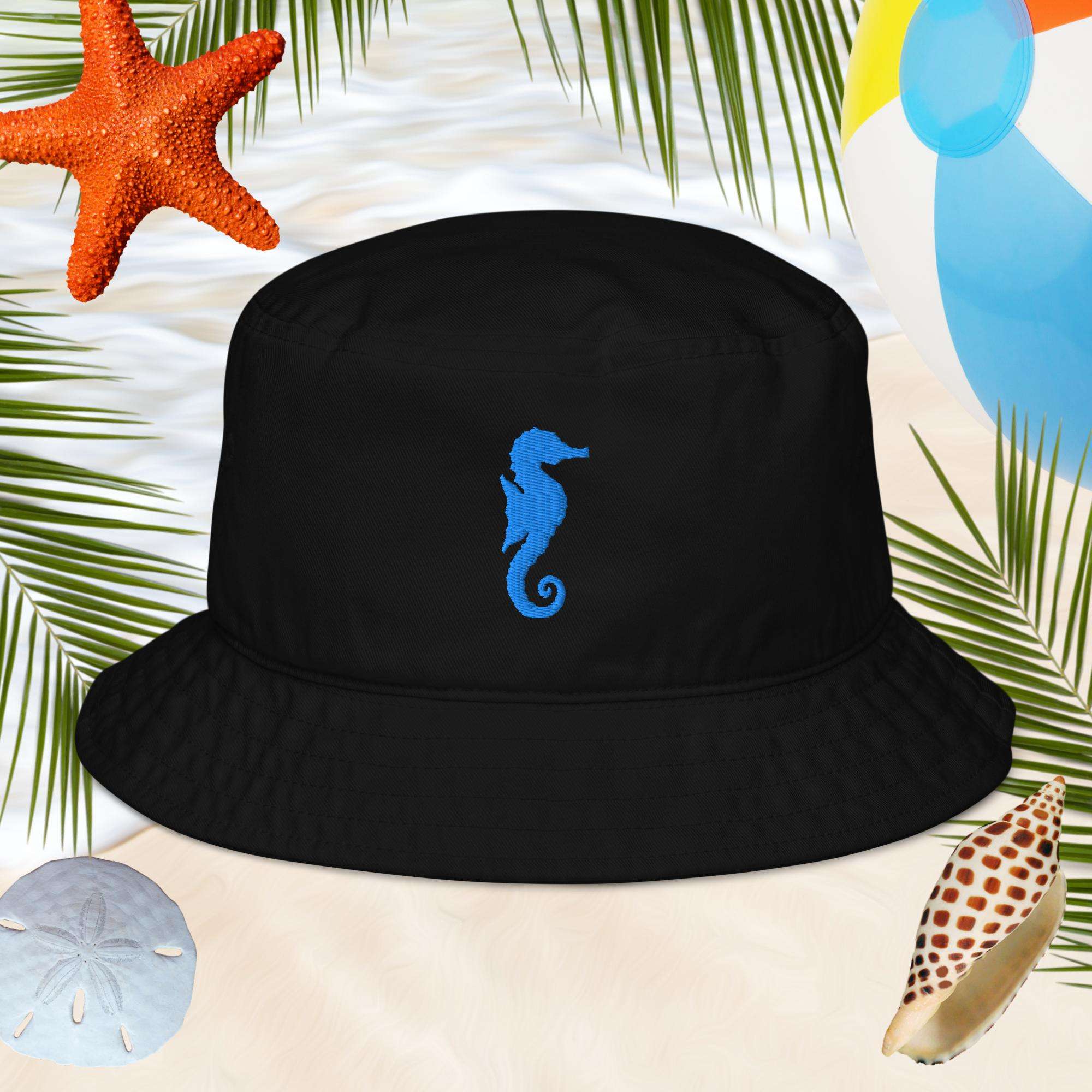 Black bucket hat with a blue seahorse embroidery on a beach-themed background with starfish, sand, and palm leaves.
