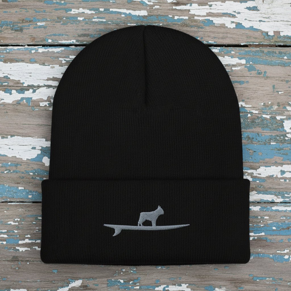A black ribbed knit beanie hat with a folded cuff, featuring a small gray embroidered graphic of a frenchie bulldog standing on a surfboard on the front.