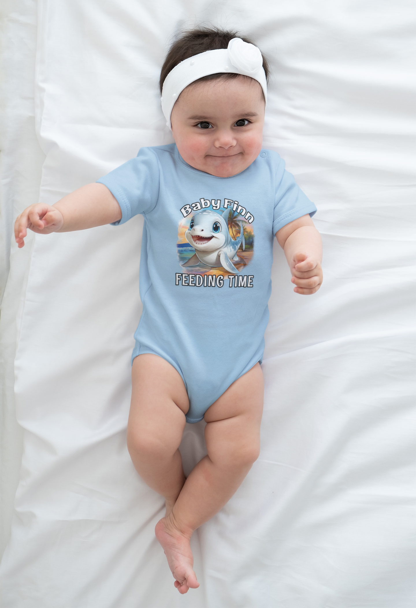 Baby wearing a light blue onesie with a cartoon baby shark and feeding time text, lying on a white blanket.