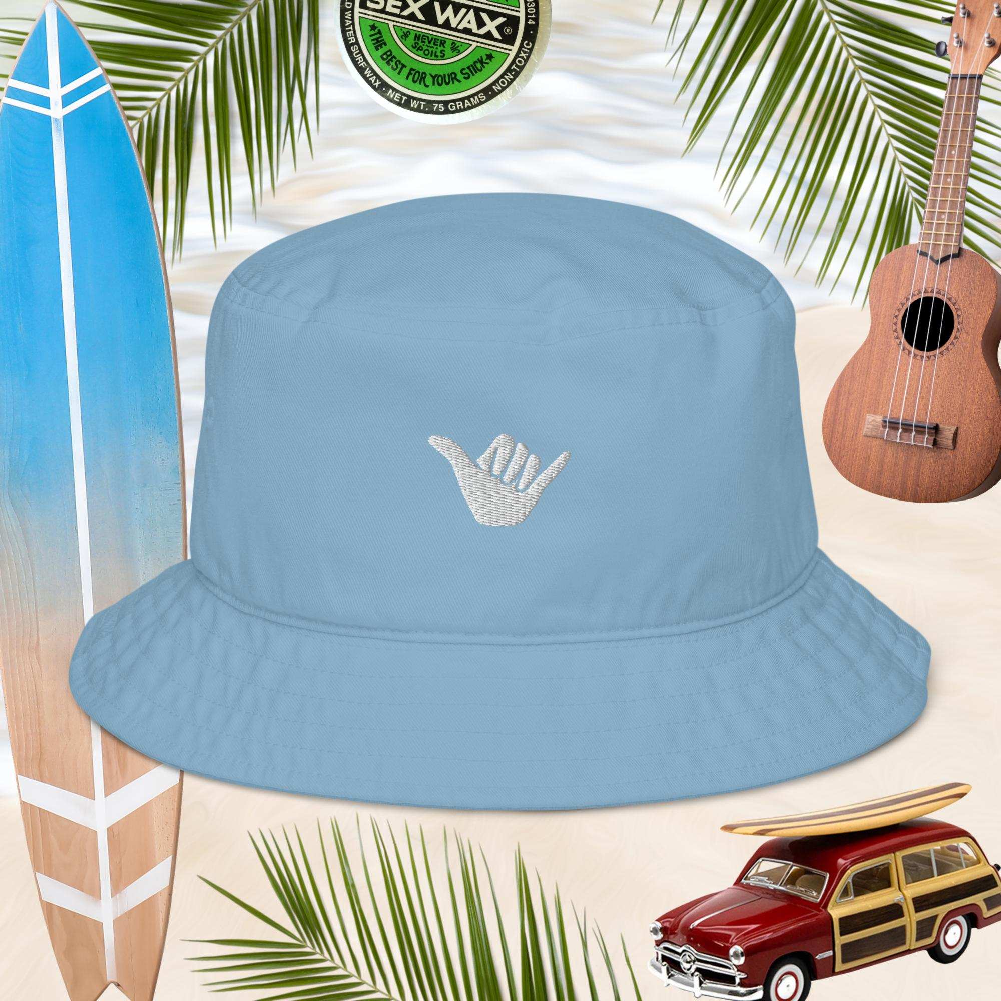 Blue bucket hat with an embroidered shaka sign on a background with a surfboard, ukulele, and vintage beach woody car.