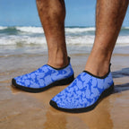 Blue patterned Blue Shark Slippers with black soles worn by a person standing on wet sand at the beach, with ocean waves and blue sky in the background.