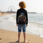 A child standing on Brighton Beach in Brooklyn, facing the ocean, wearing a black long-sleeve sun rashie with a large colorful fish graphic on the back and blue denim shorts.