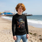 A young man with red hair stands on Brighton Beach wearing a black long-sleeve t-shirt with a colorful graphic of a spiky fish on the front, paired with light blue jeans.