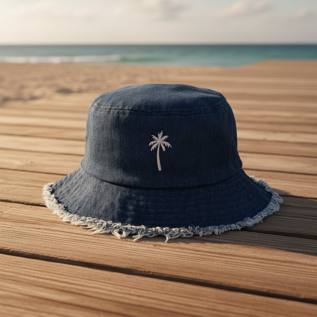 A dark blue denim bucket hat with a white embroidered palm tree on the front, resting on a wooden boardwalk with a beach and ocean in the background.