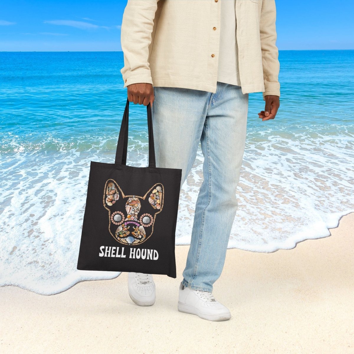A person standing on a sandy beach holds a black tote bag featuring a colorful frenchton shell-hound graphic and the text 'SHELL HOUND'. The background shows a tropical ocean scene with blue water and sky.