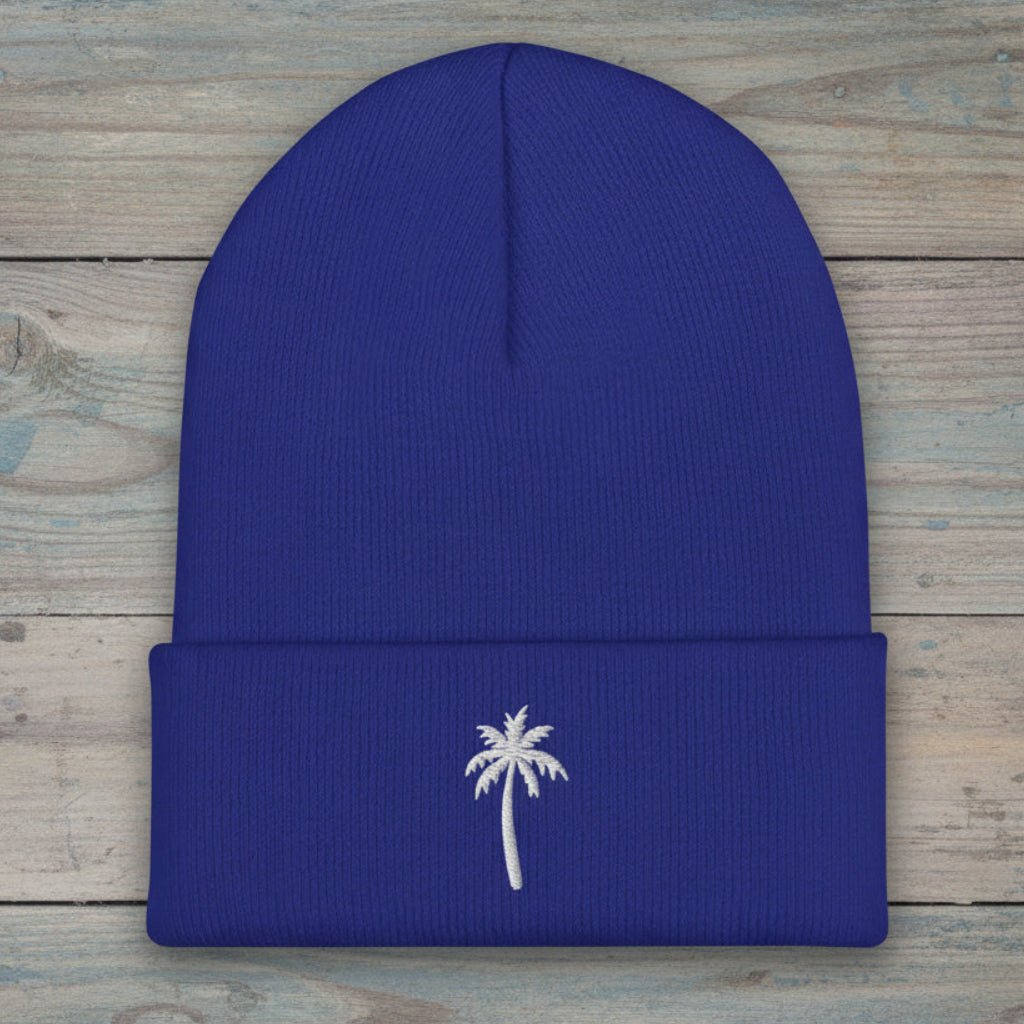 A royal blue ribbed knit beanie with a folded cuff, featuring a small white embroidered palm tree on the front, displayed flat on a rustic light blue-gray wooden plank background.