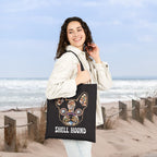 A woman standing on a sandy beach dune holds a black tote bag featuring a colorful French Bulldog illustration and the text 'SHELL HOUND'.