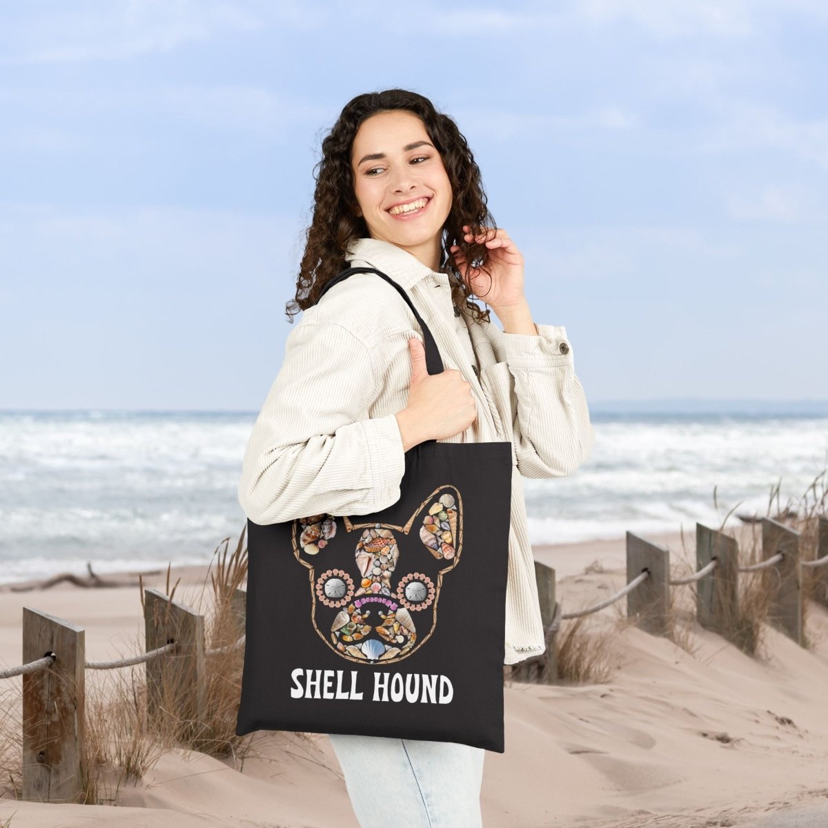 A woman standing on a sandy beach dune holds a black tote bag featuring a colorful French Bulldog illustration and the text 'SHELL HOUND'.