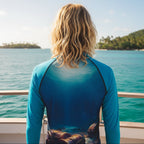 A teenage dude viewed from behind wearing a long-sleeve rashie with a blue gradient and treasure chest graphic on the lower back, standing on a boat deck with turquoise water and islands in the background.