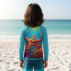 A child standing on Cayo Siesta beach facing the ocean, wearing a long-sleeve rash guard with a colorful underwater starfish graphic on the back.