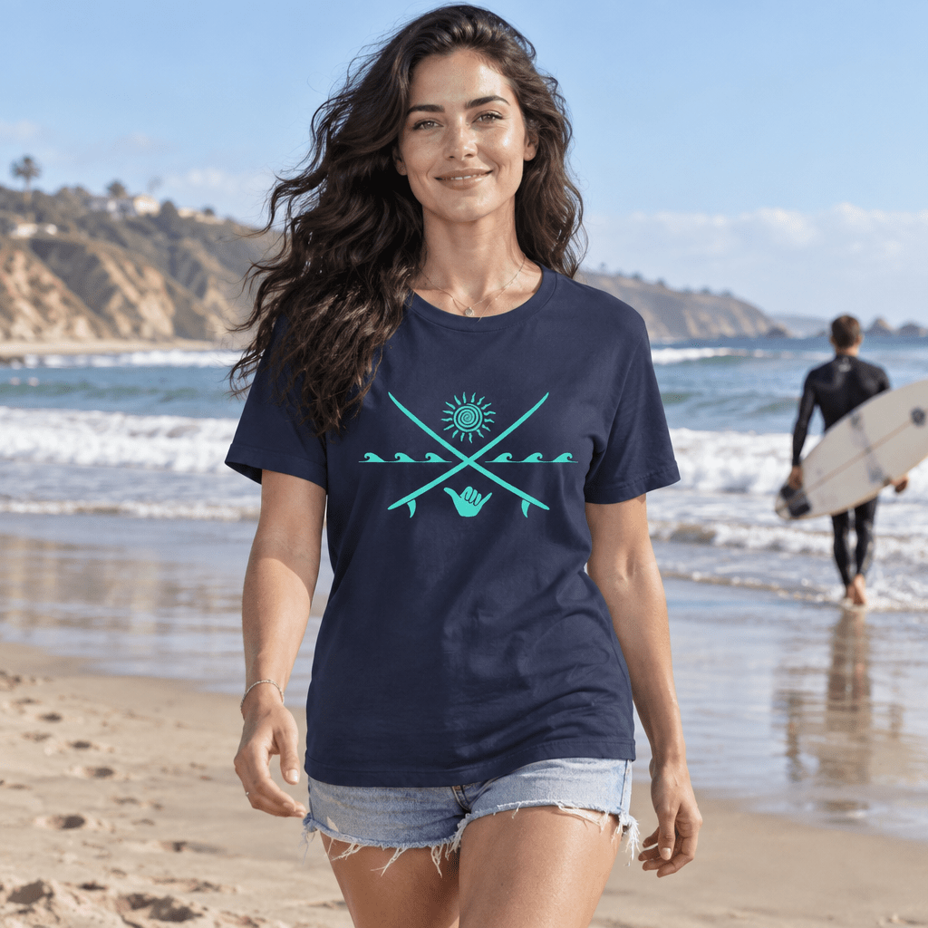 A woman walking on a sandy beach wearing a navy blue t-shirt with a turquoise graphic of crossed arrows, a sun, and a hand. A surfer is visible in the background near the ocean.