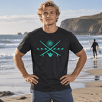 A man standing on a beach wearing a black t-shirt with a turquoise graphic of crossed swords, a sun, and a hand symbol. The background shows ocean waves, cliffs, and another surfer in the distance.