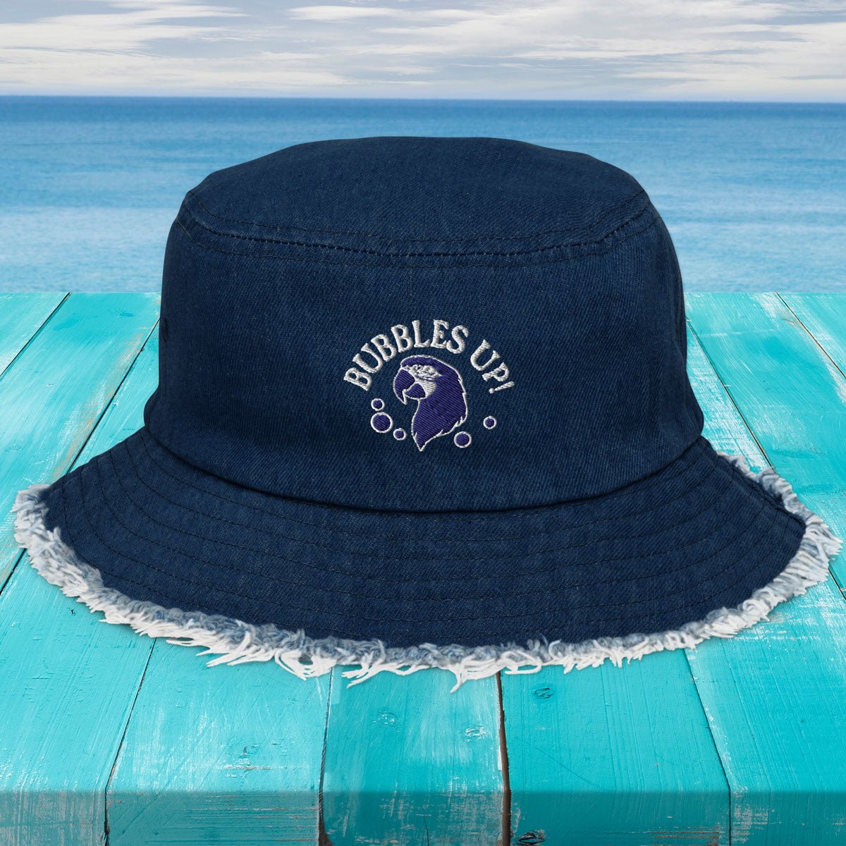 Navy bucket hat with 'Bubbles Up!' text and whale design on a turquoise wooden surface with ocean background