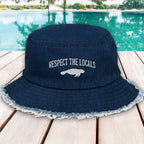 Navy blue bucket hat with 'Respect the Locals' text and manatee graphic on a wooden deck by a pool.