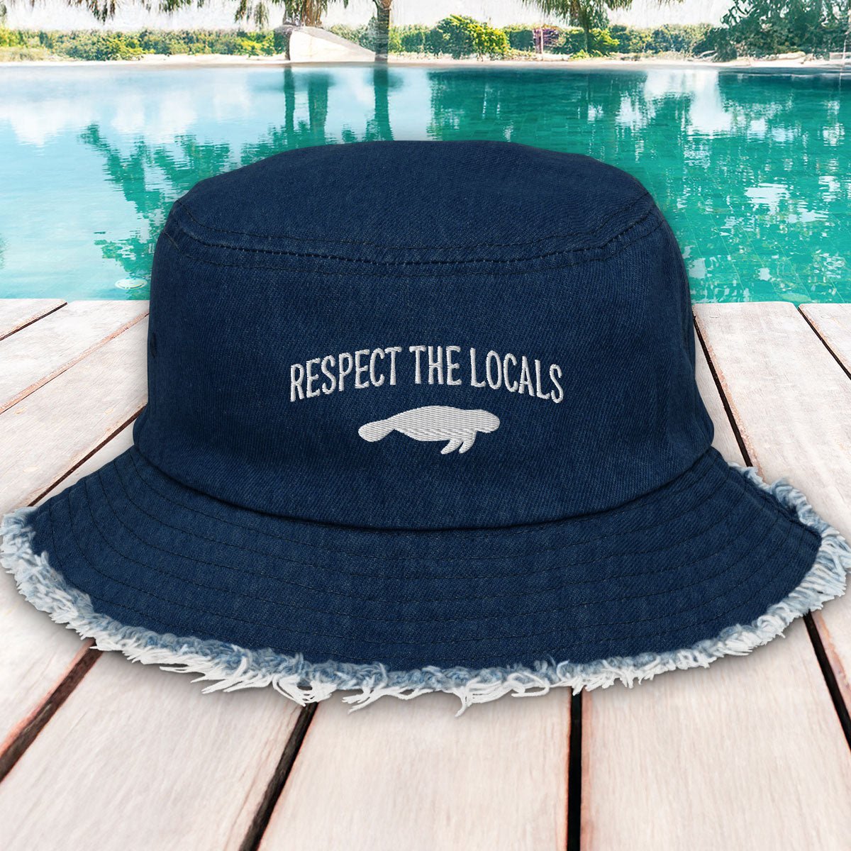 Navy blue bucket hat with 'Respect the Locals' text and manatee graphic on a wooden deck by a pool.
