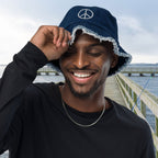 Man wearing a navy blue Denim Bucket Hat with a peace symbol, standing on a wooden pier by water.