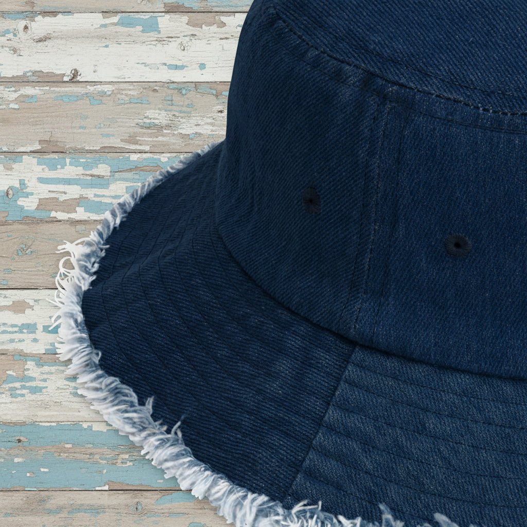 Close-up of a dark blue denim bucket hat showing the fabric texture, stitched panel seams, and a frayed, distressed edge on a nautical wood background.