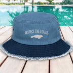 Blue bucket hat with 'Respect the Locals' text and manatee graphic on a wooden deck with pool in background