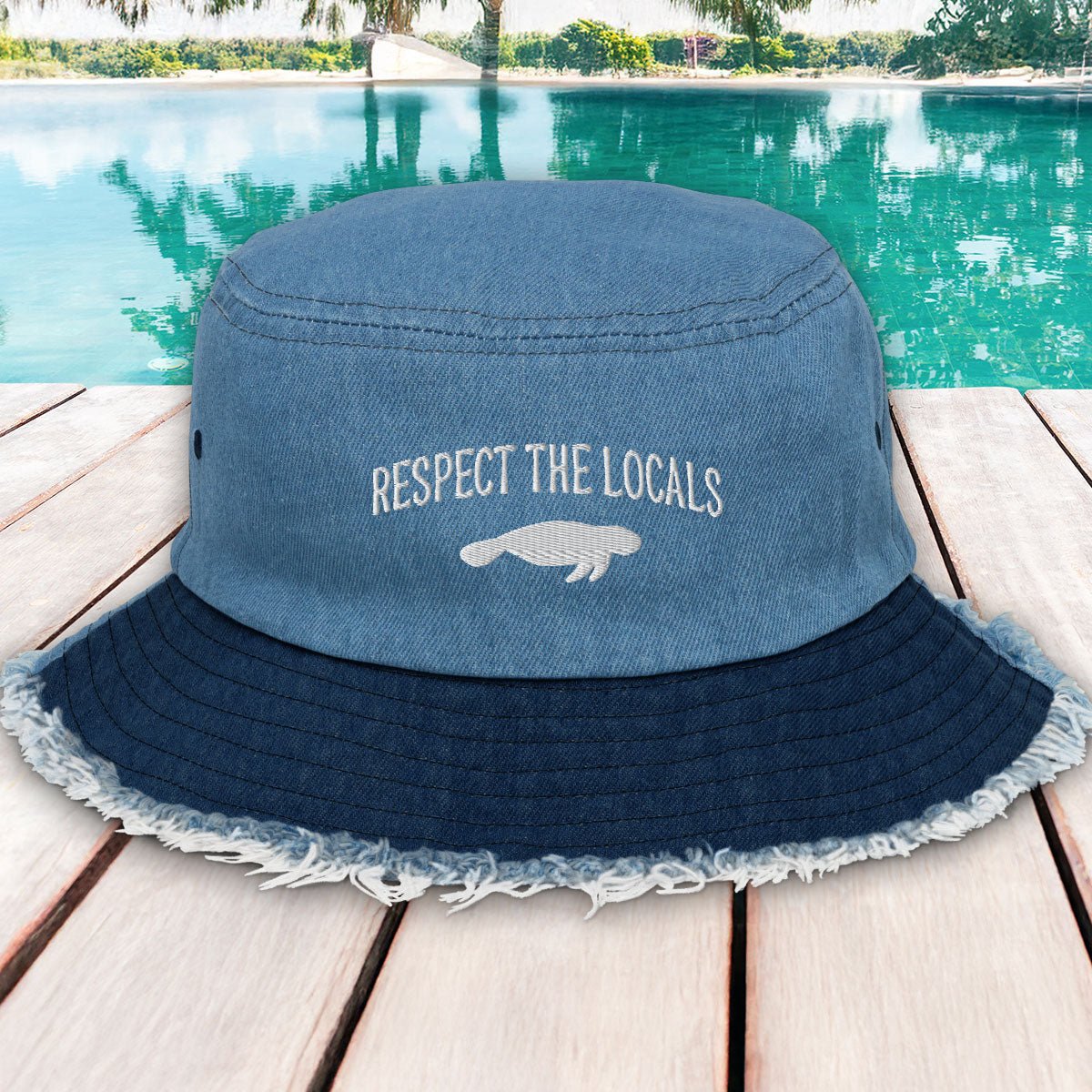 Blue bucket hat with 'Respect the Locals' text and manatee graphic on a wooden deck with pool in background