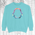 A light mint crewneck sweatshirt featuring a colorful graphic of an open mouth with sharp teeth on the chest, displayed against a white wood plank background with a palm leaf overlay.