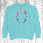 A light mint crewneck sweatshirt featuring a colorful graphic of an open mouth with sharp teeth on the chest, displayed against a white wood plank background with a palm leaf overlay.