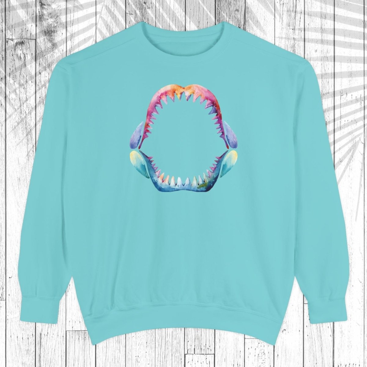A light mint crewneck sweatshirt featuring a colorful graphic of an open mouth with sharp teeth on the chest, displayed against a white wood plank background with a palm leaf overlay.