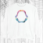 A white crewneck sweatshirt featuring a colorful, watercolor-style graphic of an open shark mouth with sharp teeth on the chest, displayed against a white wood plank background with a faint palm leaf overlay.