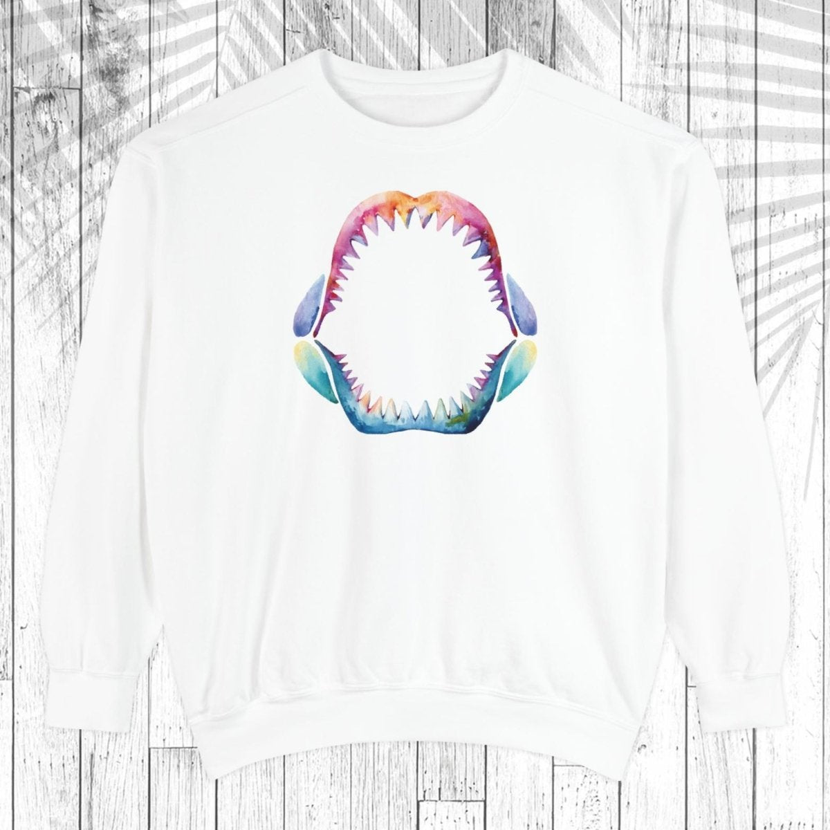 A white crewneck sweatshirt featuring a colorful, watercolor-style graphic of an open shark mouth with sharp teeth on the chest, displayed against a white wood plank background with a faint palm leaf overlay.