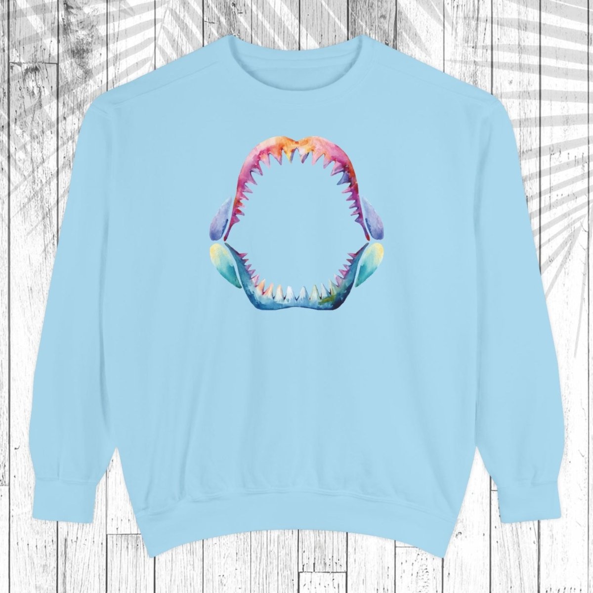 A light blue crewneck sweatshirt featuring a colorful graphic of an open shark mouth with sharp teeth on the chest, displayed against a white wood plank background with a leaf shadow overlay.