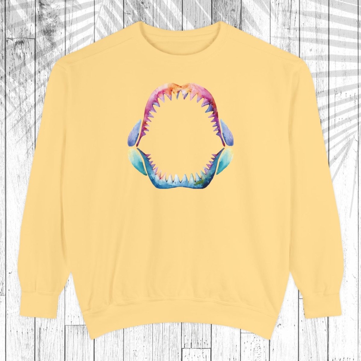 A pale yellow crewneck sweatshirt featuring a colorful graphic of an open shark mouth with sharp teeth on the chest, displayed against a white wood plank background.
