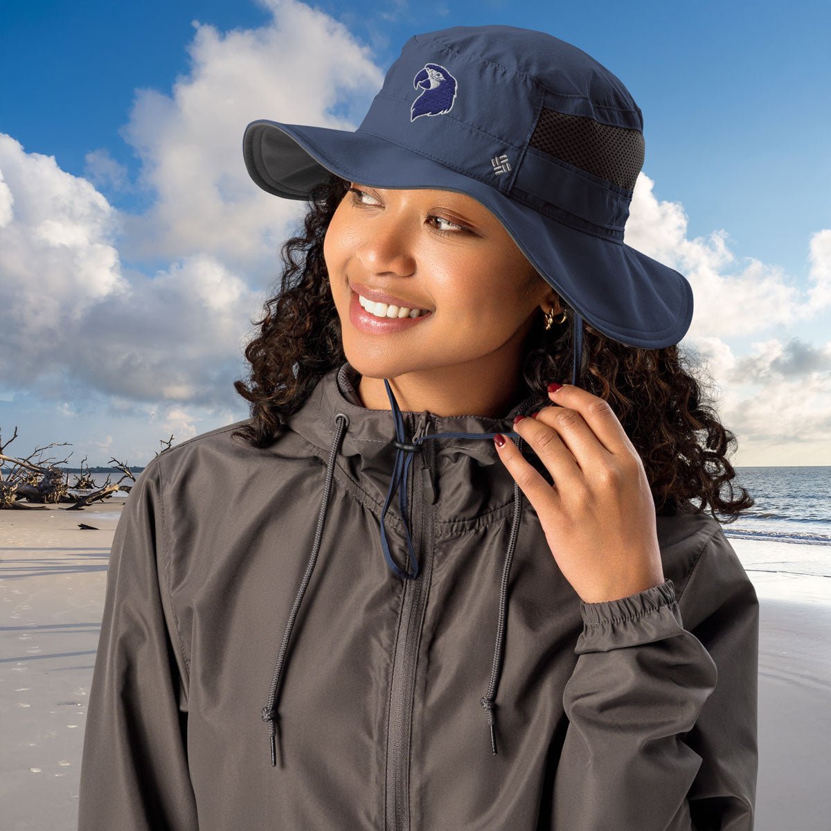 Woman wearing a navy blue Parrot Head Booney Hat and gray raincoat on a beach with ocean and sky in the background