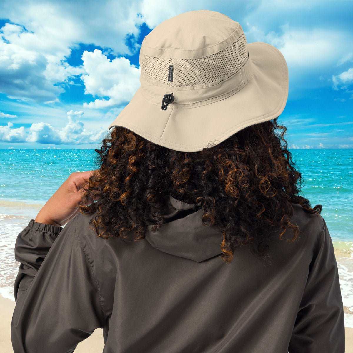 Person wearing a beige hat and dark jacket on a beach with blue sky and ocean.